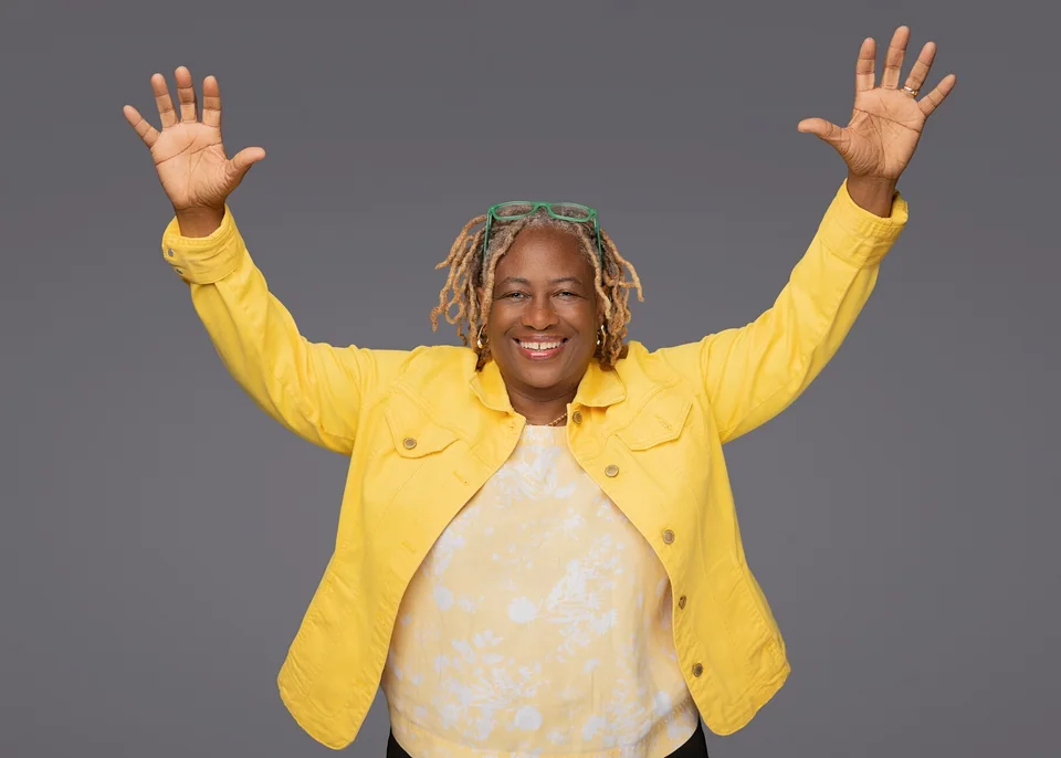 Vena Nelson, CEO of Go Big Accounting, smiling with both arms raised, in a yellow jacket over a yellow floral top, in front of a plain grey backdrop.